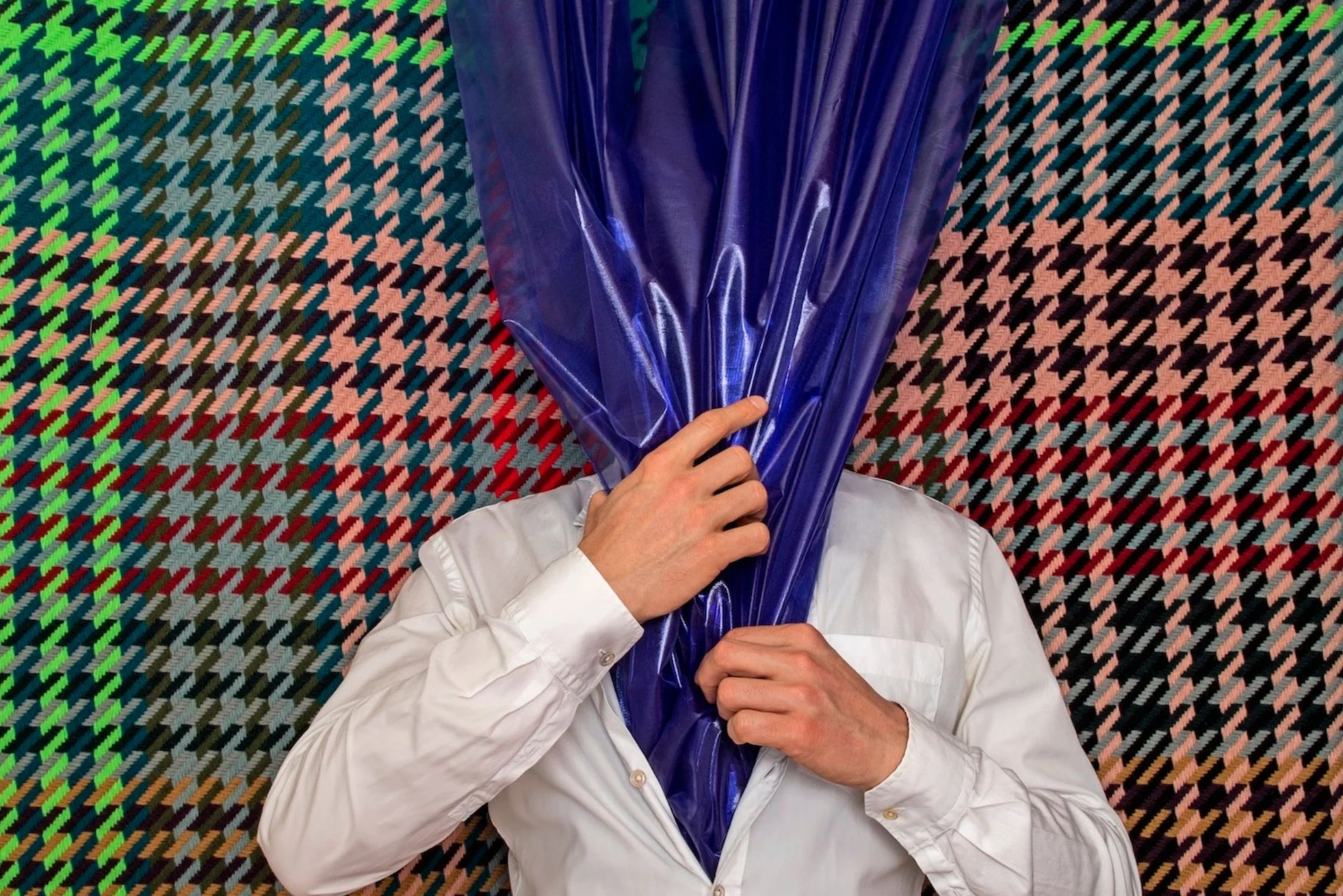 Figure in white shirt before a vivid geometric pattern with the face covered by reflective purple material